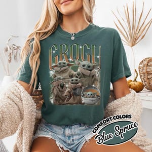 K&ouml;nnte beinhalten: Ein dunkelblau-gr&uuml;nes T-Shirt mit einem Grafikdruck der Figur Grogu in verschiedenen Posen. Der Text "GROGU" ist in einer gro&szlig;en, stilisierten Schrift &uuml;ber den Figuren abgebildet, erg&auml;nzt durch Blitzmotive und sternartige Funken im Design.