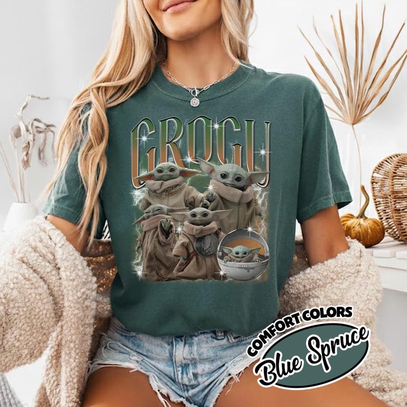 K&ouml;nnte beinhalten: Ein dunkelblau-gr&uuml;nes T-Shirt mit einem Grafikdruck der Figur Grogu in verschiedenen Posen. Der Text "GROGU" ist in einer gro&szlig;en, stilisierten Schrift &uuml;ber den Figuren abgebildet, erg&auml;nzt durch Blitzmotive und sternartige Funken im Design.