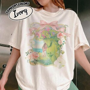 K&ouml;nnte beinhalten: Elfenbeinfarbenes T-Shirt mit floralem Design, das ein Frauengesicht, Blumen und eine rosa Schleife zeigt. Auf dem Shirt stehen die Worte "COMFORT COLORS" und "Ivory".