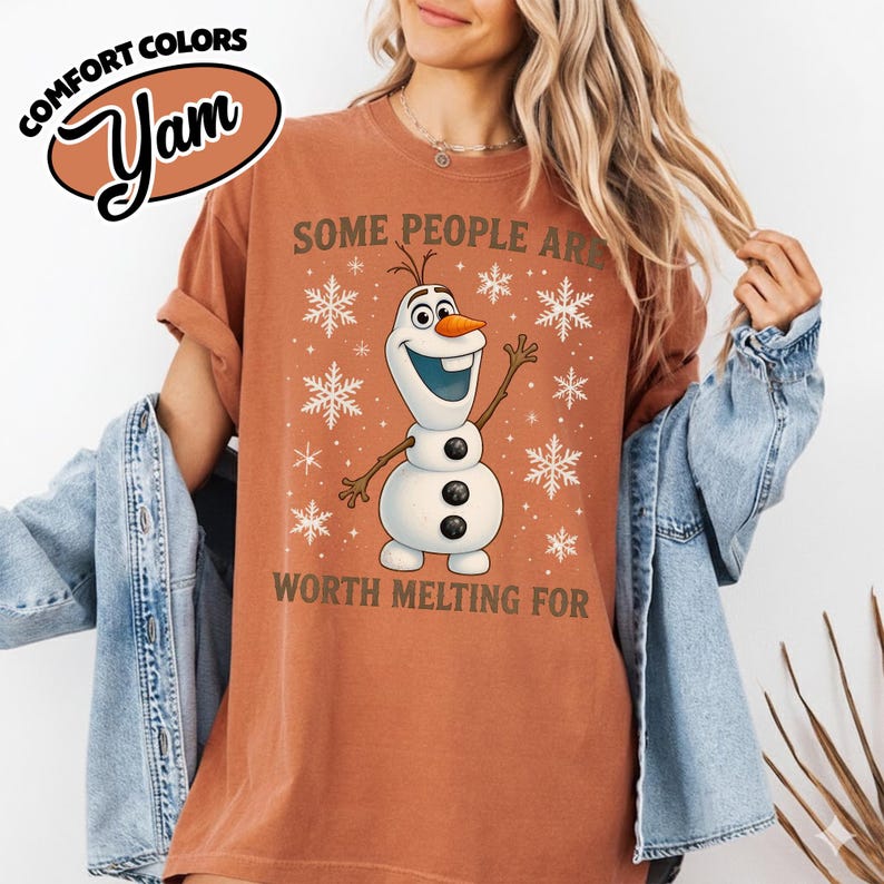 May include: A rust-colored t-shirt featuring a graphic of Olaf the snowman from Frozen, surrounded by snowflakes. The text "SOME PEOPLE ARE WORTH MELTING FOR" is printed below the character. The shirt has rolled-up sleeves.
