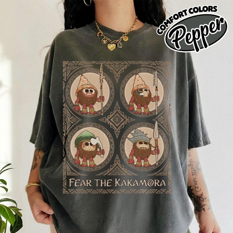May include: Dark grey t-shirt with a graphic of four cartoon characters in circular frames, each holding a spear. The text "FEAR THE KAKAMORA" is below the graphic. The shirt has the words "COMFORT COLORS Pepper" in a circular design.