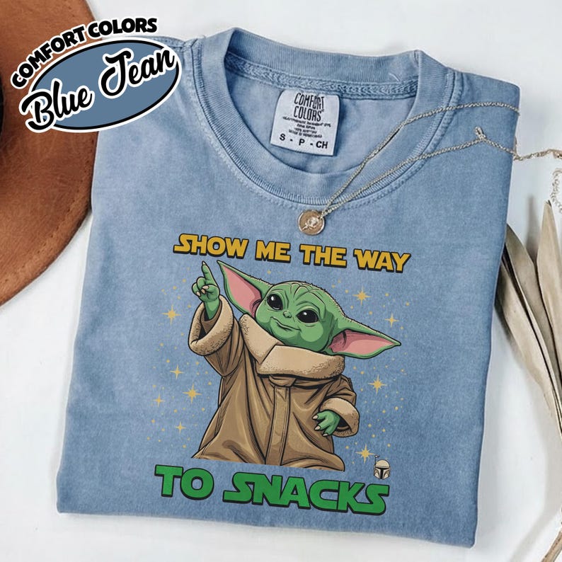 May include: Blue jean Comfort Colors t-shirt featuring a graphic of a green alien character pointing upwards, with the text "SHOW ME THE WAY TO SNACKS". The shirt also has the Comfort Colors logo and a size tag.