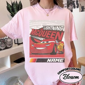 May include: A light pink t-shirt featuring a graphic print of the red race car Lightning McQueen. The design includes the text LIGHTNING MCQUEEN, the number 95, and a placeholder area labeled NAME. The shirt has a crew neckline and short sleeves.