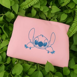 May include: A light pink tote bag featuring a blue cartoon character design. The character has large ears, big eyes, and a friendly expression. The bag is rectangular and appears to be made of a soft material. The background is a bed of green leaves.