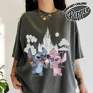 Puede incluir: Camiseta gris oscuro con una ilustraci&oacute;n de dibujos animados de Stitch y Angel sosteniendo conos de helado frente a un castillo. La camiseta tiene las palabras "COMFORT COLORS" y "Pepper" en un dise&ntilde;o circular.