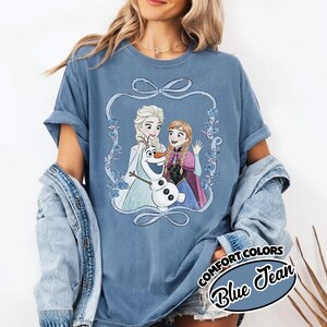 May include: Blue t-shirt featuring a graphic of Elsa, Anna, and Olaf from Frozen, framed by a light blue ribbon and floral design. The shirt has rolled-up sleeves and the words "COMFORT COLORS Blue Jean" in a circular design.