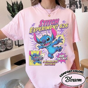 Może przedstawiać: Jasnor&oacute;żowy t-shirt z grafiką przedstawiającą niebieską postać Stitcha z r&oacute;żowymi uszami. Tekst głosi Stitch Experiment 626, Hear the Call oraz A Complete Mayhem. Mniejszy kwadratowy panel pokazuje Stitcha trzymającego kolorową broń laserową.