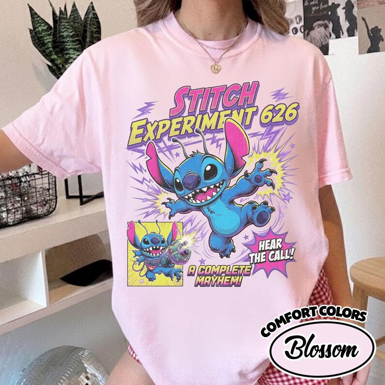Może przedstawiać: Jasnor&oacute;żowy t-shirt z grafiką przedstawiającą niebieską postać Stitcha z r&oacute;żowymi uszami. Tekst głosi Stitch Experiment 626, Hear the Call oraz A Complete Mayhem. Mniejszy kwadratowy panel pokazuje Stitcha trzymającego kolorową broń laserową.