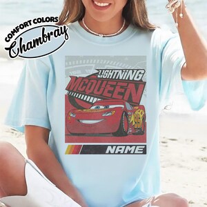 May include: A light blue t-shirt featuring a graphic of the red race car Lightning McQueen with the text Lightning McQueen and Name. The shirt includes a Comfort Colors Chambray label in the upper left corner and a retro speedometer design.