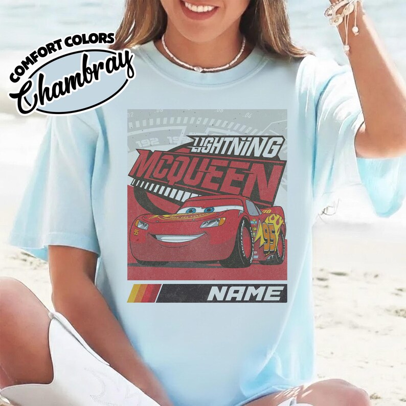 May include: A light blue t-shirt featuring a graphic of the red race car Lightning McQueen with the text Lightning McQueen and Name. The shirt includes a Comfort Colors Chambray label in the upper left corner and a retro speedometer design.
