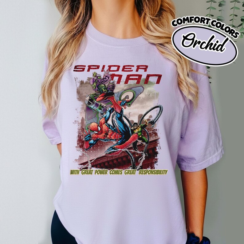 Op de afbeelding: Orchid Comfort Colors T-shirt met een Spider-Man afbeelding. Het ontwerp toont Spider-Man in actie met de tekst "SPIDER-MAN" en de zin "WITH GREAT POWER COMES GREAT RESPONSIBILITY".