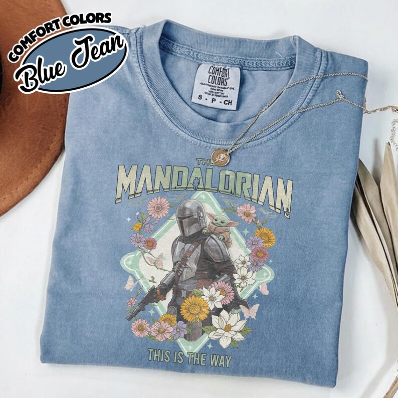 May include: A blue jean colored Comfort Colors t-shirt featuring a graphic of The Mandalorian and Grogu surrounded by colorful flowers and butterflies. The text reads The Mandalorian and This Is The Way. A silver necklace rests on the collar.