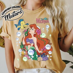 K&ouml;nnte beinhalten: Ein senfgelbes T-Shirt mit einer Grafik einer rothaarigen Meerjungfrau und eines gelb-blauen Fisches, umgeben von Blasen, Korallen und Blumen. Der Text "THE LITTLE MERMAID" ist in Pink dar&uuml;ber gedruckt, inklusive eines "COMFORT COLORS MUSTARD"-Etiketts.