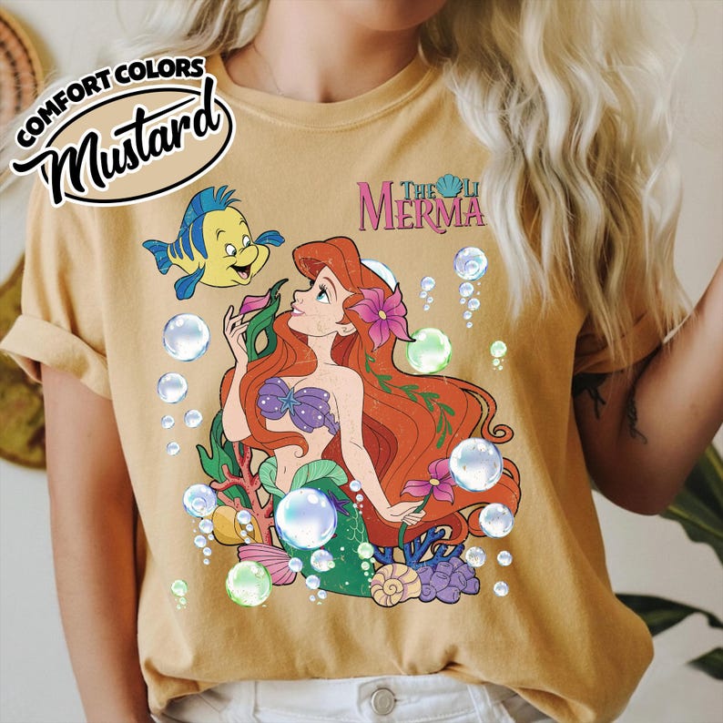 K&ouml;nnte beinhalten: Ein senfgelbes T-Shirt mit einer Grafik einer rothaarigen Meerjungfrau und eines gelb-blauen Fisches, umgeben von Blasen, Korallen und Blumen. Der Text "THE LITTLE MERMAID" ist in Pink dar&uuml;ber gedruckt, inklusive eines "COMFORT COLORS MUSTARD"-Etiketts.