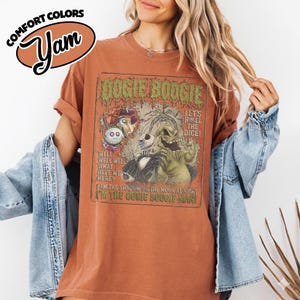 May include: Rust-colored t-shirt with a graphic featuring the text "Oogie Boogie" and various characters. The shirt also includes the text "Let's Roll the Dice!" and "I'm the Oogie Boogie Man!" The shirt has the "Comfort Colors" and "Yam" logos.