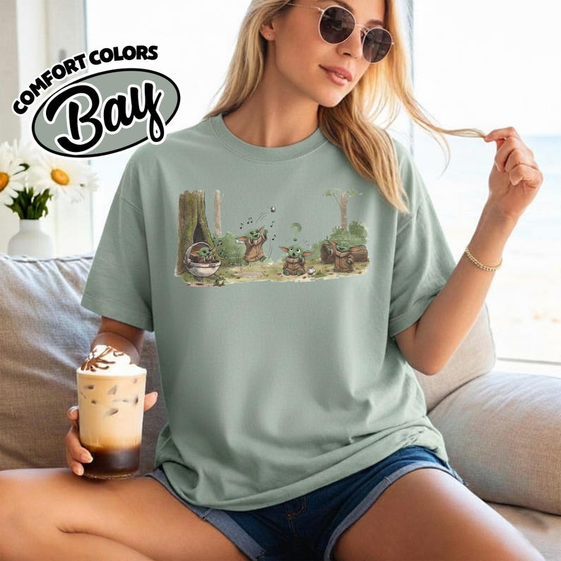 May include: A light green Comfort Colors Bay t-shirt featuring a graphic of Grogu from The Mandalorian. The illustration shows Grogu in various poses in a forest setting. The shirt has a relaxed fit.
