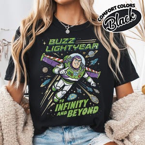 May include: A black t-shirt featuring a graphic of Buzz Lightyear in a space suit with purple wings. The text reads BUZZ LIGHTYEAR at the top and TO INFINITY AND BEYOND at the bottom, surrounded by planets, stars, and small spaceships in space.