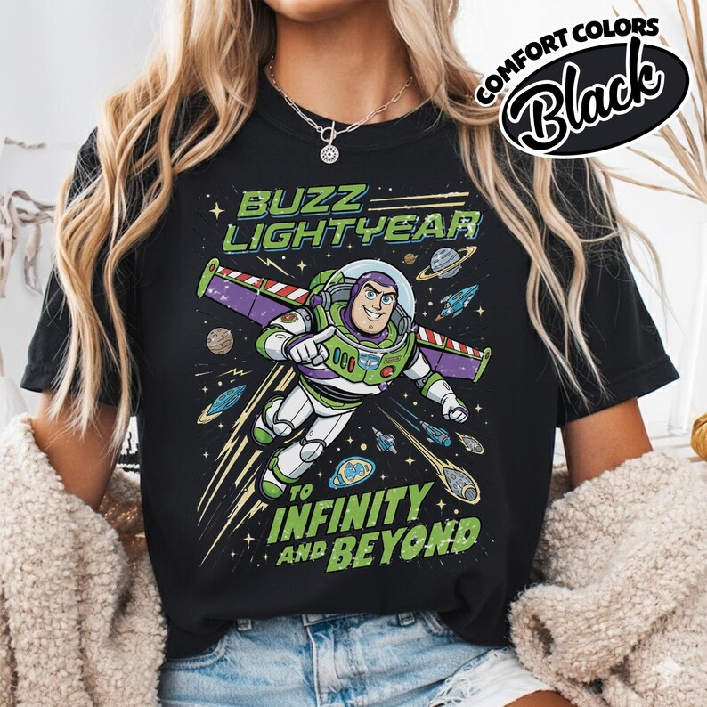 May include: A black t-shirt featuring a graphic of Buzz Lightyear in a space suit with purple wings. The text reads BUZZ LIGHTYEAR at the top and TO INFINITY AND BEYOND at the bottom, surrounded by planets, stars, and small spaceships in space.