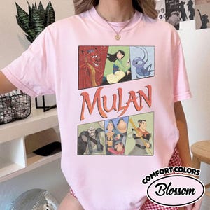 Puede incluir: Una camiseta Comfort Colors rosa claro con un gr&aacute;fico de la pel&iacute;cula de Disney Mulan. El dise&ntilde;o incluye la palabra "Mulan" en letras naranjas y paneles de personajes. La camiseta tambi&eacute;n tiene la palabra "Blossom" en un &oacute;valo blanco.