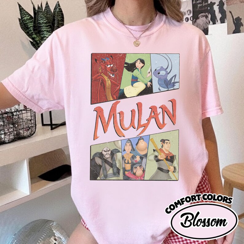 Puede incluir: Una camiseta Comfort Colors rosa claro con un gr&aacute;fico de la pel&iacute;cula de Disney Mulan. El dise&ntilde;o incluye la palabra "Mulan" en letras naranjas y paneles de personajes. La camiseta tambi&eacute;n tiene la palabra "Blossom" en un &oacute;valo blanco.