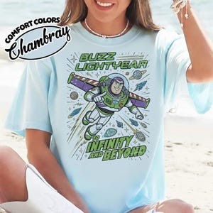 May include: A light blue t-shirt featuring a graphic of Buzz Lightyear flying through space with the text BUZZ LIGHTYEAR TO INFINITY AND BEYOND. The design includes planets, stars, and a rocket ship. The shirt has a Comfort Colors Chambray label.