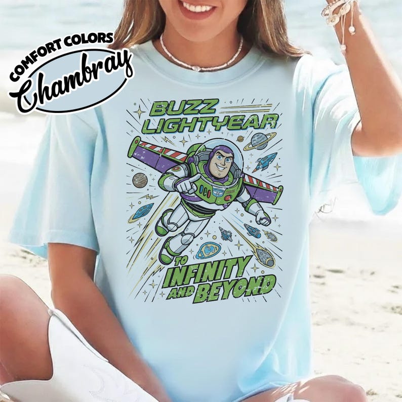 May include: A light blue t-shirt featuring a graphic of Buzz Lightyear flying through space with the text BUZZ LIGHTYEAR TO INFINITY AND BEYOND. The design includes planets, stars, and a rocket ship. The shirt has a Comfort Colors Chambray label.