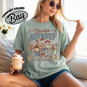 May include: Sage green t-shirt featuring the text "Woody's Roundup" with a cartoon graphic of Woody, Jessie, and other characters. The shirt also has the text "You've Got A Friend In Me!" and the Comfort Colors Bay logo.