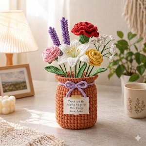 May include: A crocheted flower arrangement in an orange vase. The bouquet includes red, white, pink, and yellow flowers, with green leaves and purple accents. A small tag on the vase reads "Thank you for helping me grow Mrs. Emily Love, Anna."