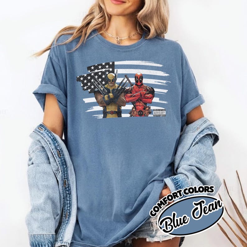 May include: A blue jean colored t-shirt featuring a graphic print of Wolverine and Deadpool standing in front of a stylized American flag. The design includes a small black rectangular box with the text ADVISORY EXPLICIT CONTENT.