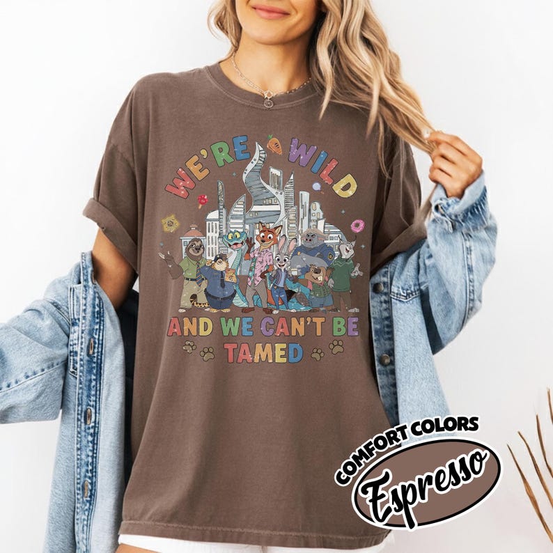 May include: Brown t-shirt with a colorful graphic featuring cartoon characters and the text "WE'RE WILD AND WE CAN'T BE TAMED." The design includes a cityscape and paw prints. The shirt is labeled "COMFORT COLORS Espresso."
