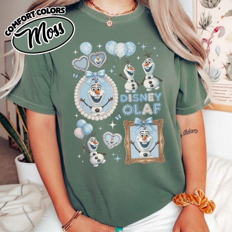 May include: Moss green t-shirt featuring the text "Disney Olaf" with images of Olaf the snowman, balloons, and hearts. The shirt has a round neck and short sleeves. The Comfort Colors logo is in the upper left corner.