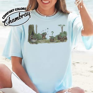 May include: Light blue t-shirt with a graphic of Baby Yoda characters in a forest scene. The shirt has the words "COMFORT COLORS" and "Chambray" printed on the upper left side. The shirt is made of a soft material.