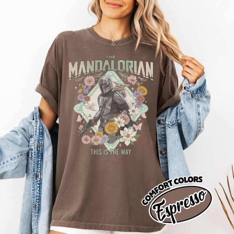 May include: A brown short-sleeve t-shirt featuring a graphic print of The Mandalorian and Grogu surrounded by colorful flowers and butterflies. The text reads THE MANDALORIAN and THIS IS THE WAY. A label indicates the color is Comfort Colors Espresso.