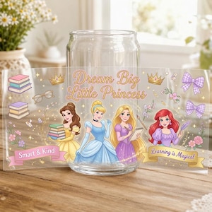 May include: A clear glass tumbler featuring a wrap-around design with illustrations of four princesses, stacks of books, bows, and crowns. The text reads Dream Big Little Princess, Smart & Kind, and Learning is Magical in gold and pink script.