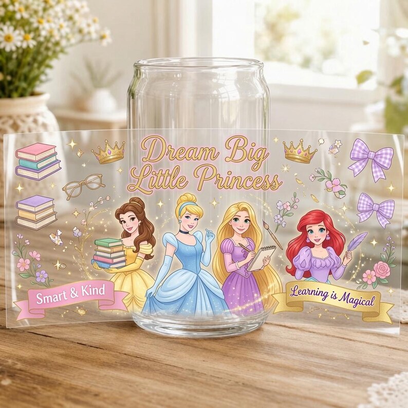 May include: A clear glass tumbler featuring a wrap-around design with illustrations of four princesses, stacks of books, bows, and crowns. The text reads Dream Big Little Princess, Smart & Kind, and Learning is Magical in gold and pink script.