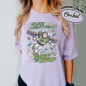 May include: A light purple t-shirt featuring a graphic of Buzz Lightyear flying through space with planets and stars. The text reads BUZZ LIGHTYEAR TO INFINITY AND BEYOND. A small oval badge in the corner displays the text COMFORT COLORS Orchid.