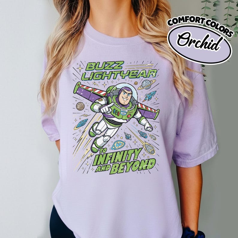 May include: A light purple t-shirt featuring a graphic of Buzz Lightyear flying through space with planets and stars. The text reads BUZZ LIGHTYEAR TO INFINITY AND BEYOND. A small oval badge in the corner displays the text COMFORT COLORS Orchid.