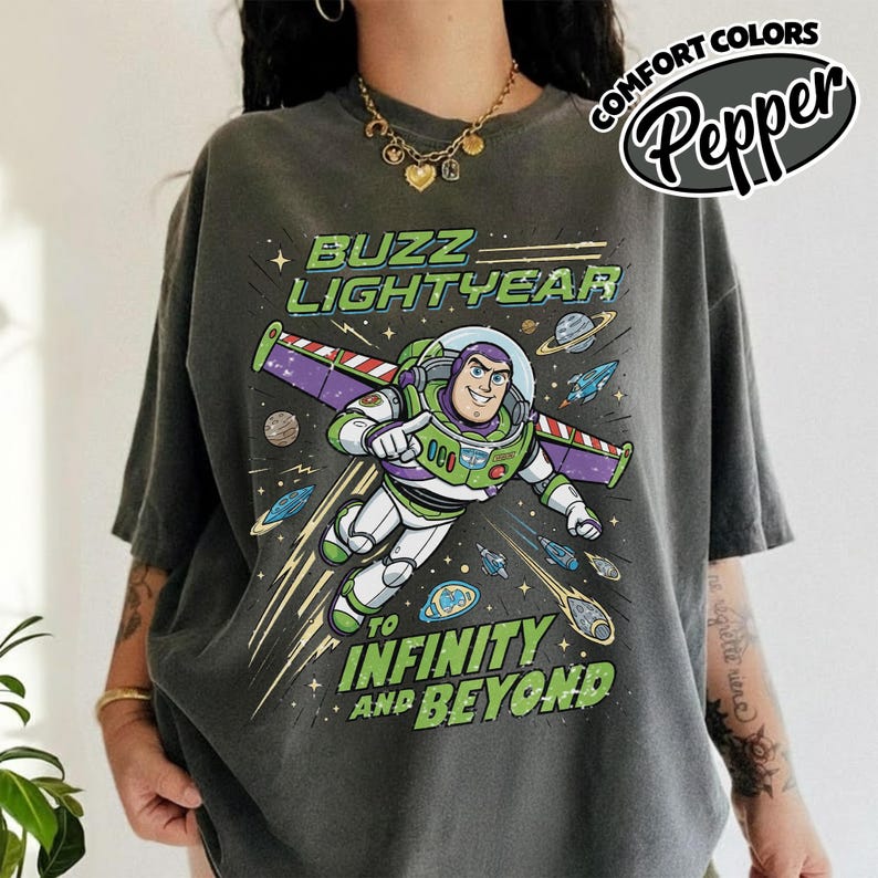May include: A charcoal grey short-sleeve t-shirt featuring a graphic of Buzz Lightyear flying through space. The text reads BUZZ LIGHTYEAR TO INFINITY AND BEYOND. The design includes planets, stars, and small spacecraft against a dark background.