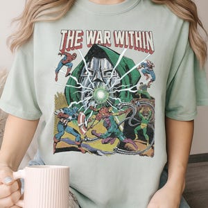 May include: A light green t-shirt featuring a comic book-style graphic. The graphic includes the text "THE WAR WITHIN" in red, along with illustrations of several superheroes and a villain. The design is reminiscent of vintage comic book art.