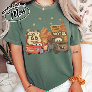 May include: Moss green t-shirt featuring a graphic of Lightning McQueen and Mater from the movie Cars, with a Route 66 sign and Cozy Cone Motel. The shirt has the words "Comfort Colors" and "Moss" in a circular design.