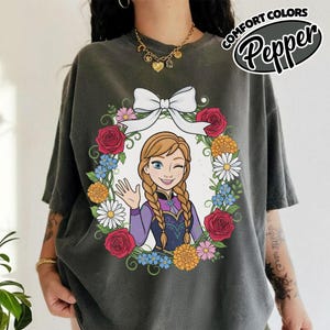 Peut inclure: T-shirt gris fonc&eacute; avec une image de dessin anim&eacute; d'une femme faisant un clin d'&oelig;il et saluant, entour&eacute;e d'une couronne florale et d'un n&oelig;ud blanc. Le texte "COMFORT COLORS Pepper" est imprim&eacute; en haut &agrave; droite.