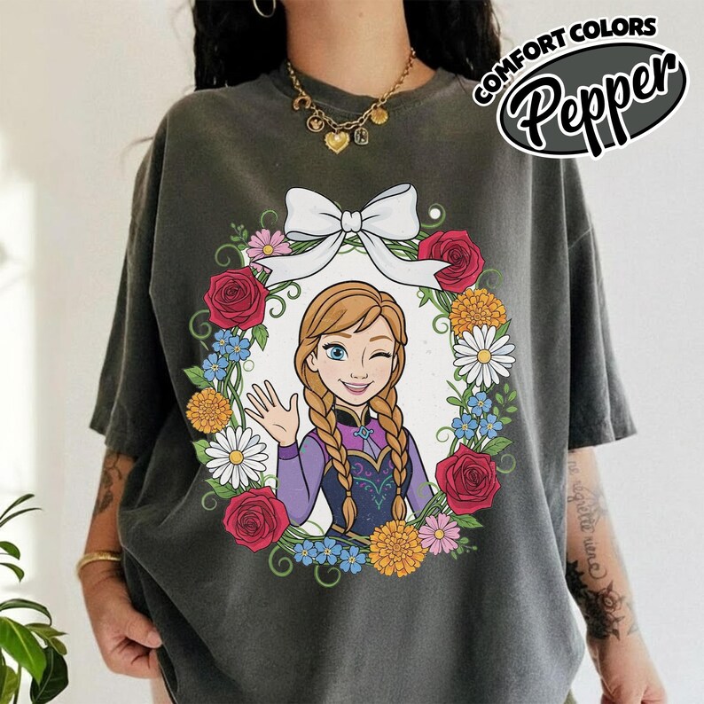 Peut inclure: T-shirt gris fonc&eacute; avec une image de dessin anim&eacute; d'une femme faisant un clin d'&oelig;il et saluant, entour&eacute;e d'une couronne florale et d'un n&oelig;ud blanc. Le texte "COMFORT COLORS Pepper" est imprim&eacute; en haut &agrave; droite.
