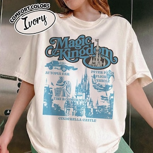 May include: Ivory-colored t-shirt with a blue graphic featuring the words "Magic Kingdom" and illustrations of Autopia Car, Peter Pan's Flight Vehicle, Tiki Room Drummer, and Cinderella Castle. The shirt is labeled "Comfort Colors."