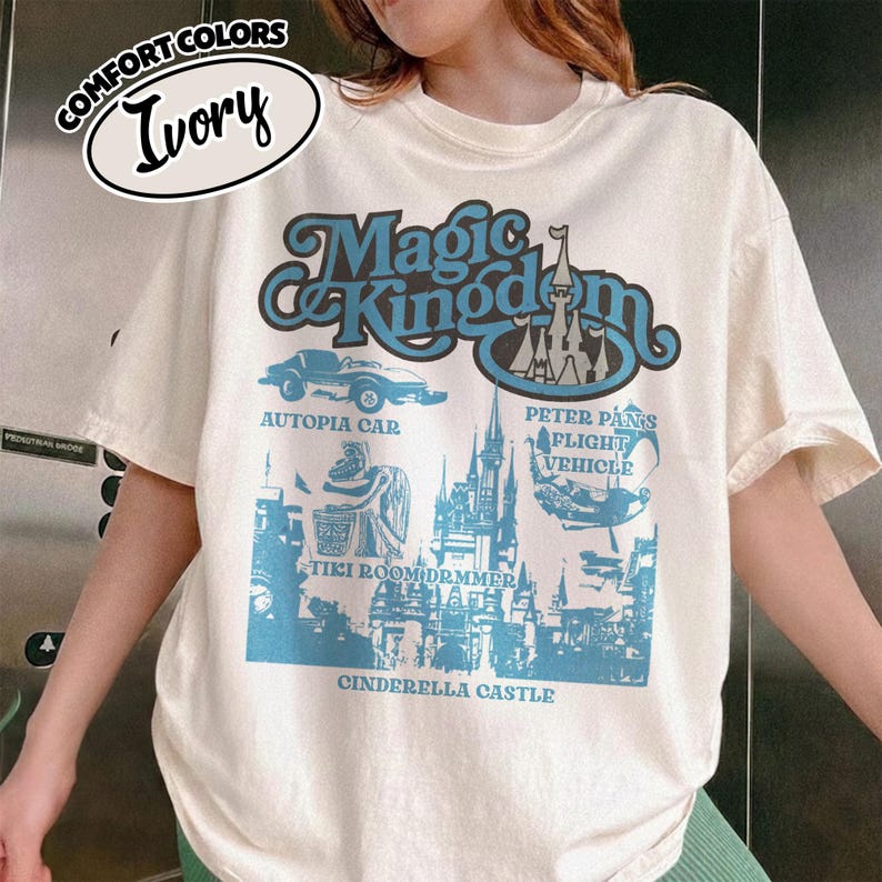 May include: Ivory-colored t-shirt with a blue graphic featuring the words "Magic Kingdom" and illustrations of Autopia Car, Peter Pan's Flight Vehicle, Tiki Room Drummer, and Cinderella Castle. The shirt is labeled "Comfort Colors."