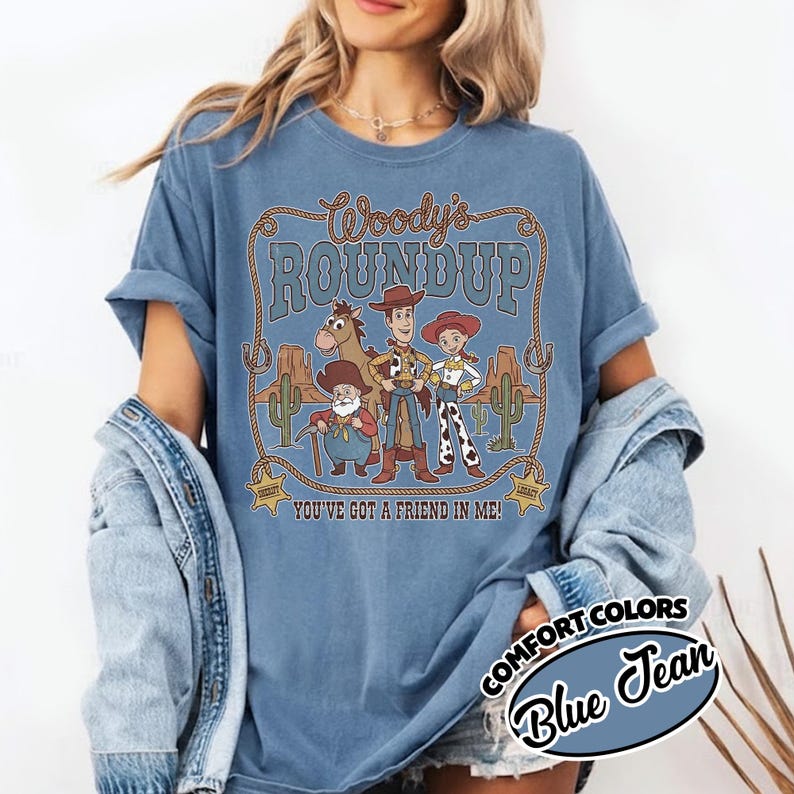 May include: Blue Comfort Colors t-shirt featuring a graphic of Woody's Roundup characters. The design includes the text "Woody's Roundup" and "You've Got a Friend in Me!" in a western-themed frame. The shirt is labeled "Blue Jean".