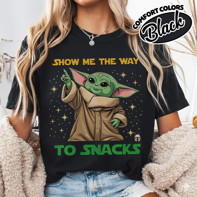 May include: Black t-shirt featuring a graphic of a green alien character with large ears, pointing upwards. The text "SHOW ME THE WAY TO SNACKS" is printed in yellow and green. The shirt has a Comfort Colors label.
