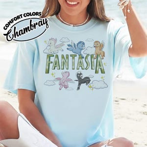 May include: Pale blue t-shirt featuring the word "Fantasia" in green, with cartoon pegasus figures. The shirt also has the words "Comfort Colors Chambray" in a circular design.