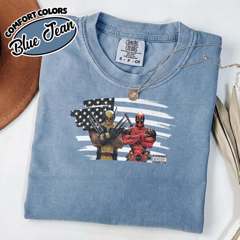 May include: A blue jean colored t-shirt featuring a graphic of Wolverine and Deadpool standing in front of a stylized American flag. The shirt has a Comfort Colors label at the neck and a small Parental Advisory label printed near the graphic.