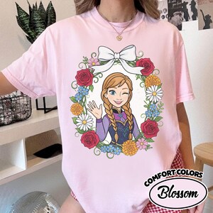 Peut inclure: T-shirt rose Comfort Colors avec une illustration de dessin anim&eacute; d'un personnage aux tresses blondes, faisant un clin d'&oelig;il et saluant, entour&eacute; d'une couronne florale et d'un n&oelig;ud blanc. Le texte "Comfort Colors Blossom" est imprim&eacute; en bas &agrave; droite.