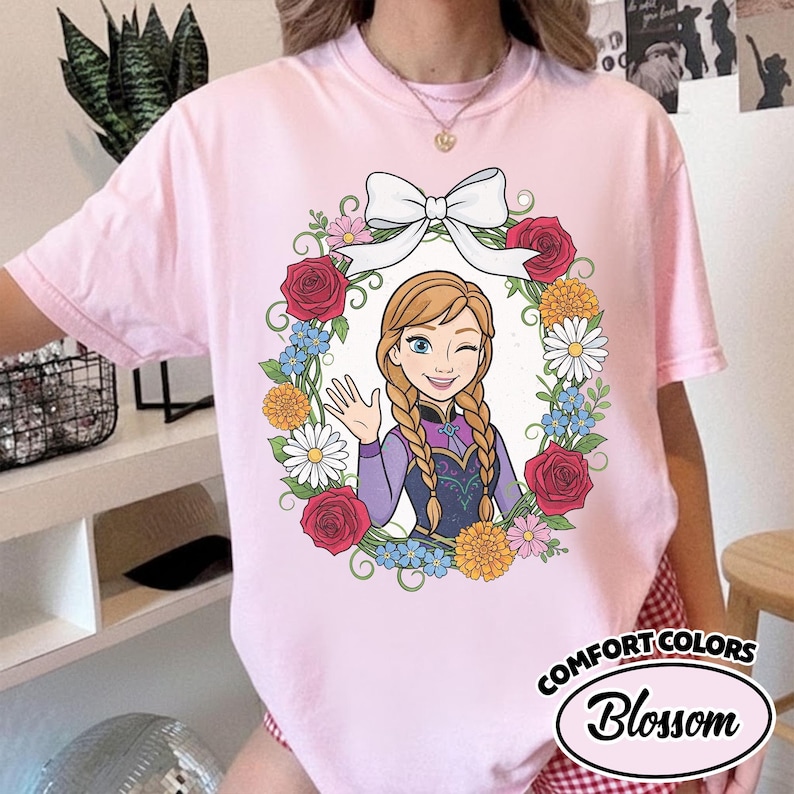 Peut inclure: T-shirt rose Comfort Colors avec une illustration de dessin anim&eacute; d'un personnage aux tresses blondes, faisant un clin d'&oelig;il et saluant, entour&eacute; d'une couronne florale et d'un n&oelig;ud blanc. Le texte "Comfort Colors Blossom" est imprim&eacute; en bas &agrave; droite.
